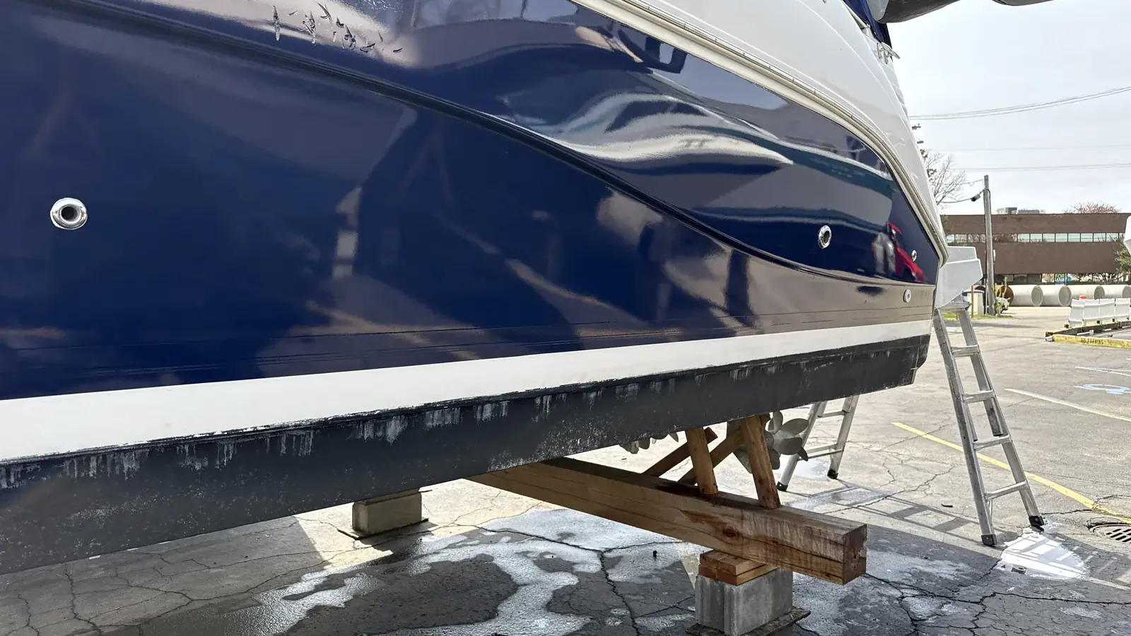 The hull of a navy cabin cruiser on blocks with a ladder leaning against it
