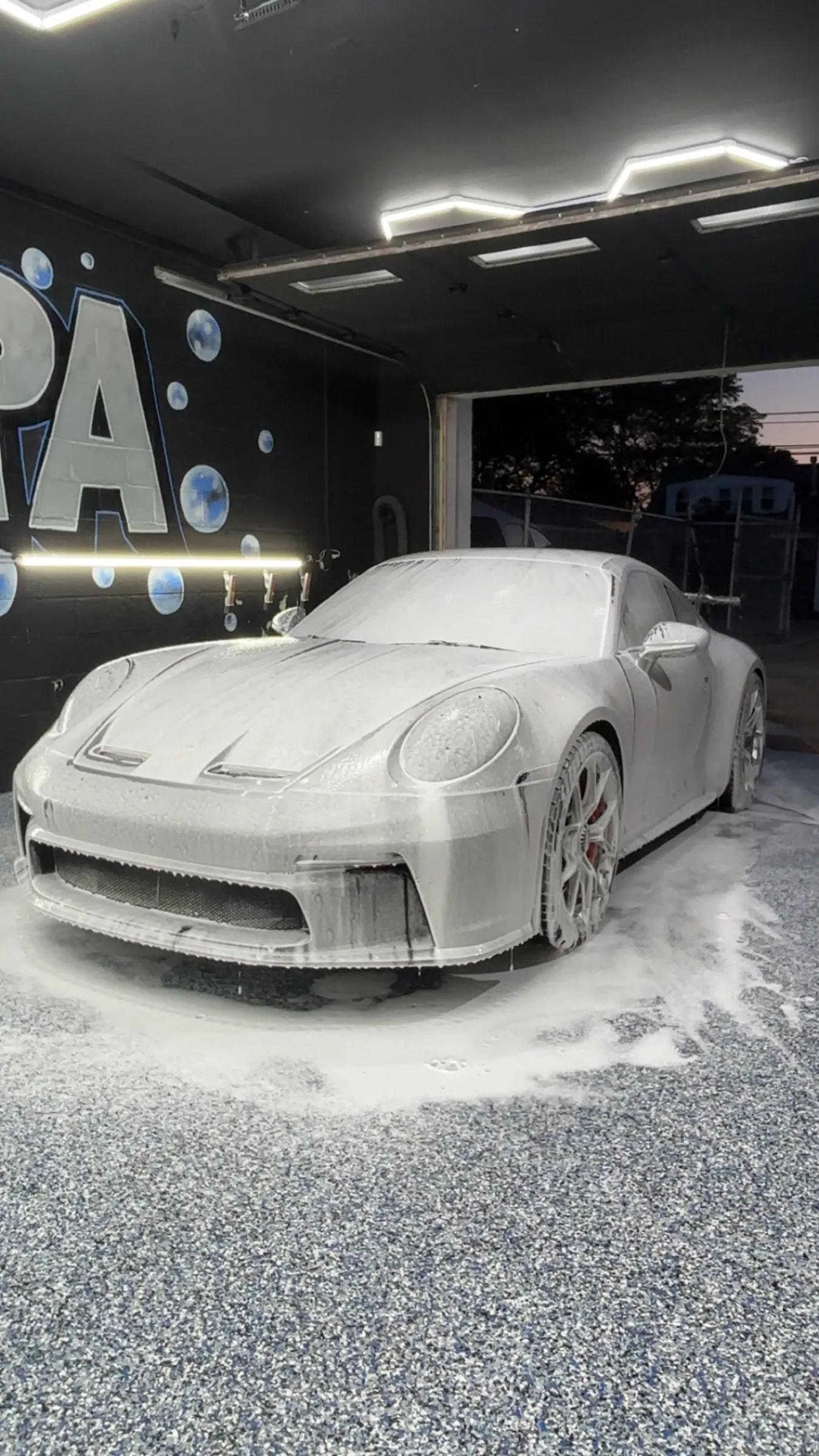 A Porsche 911 GT3 covered in snow foam under the hexagonal ceiling lights inside the Bubbles Auto Spa studio
